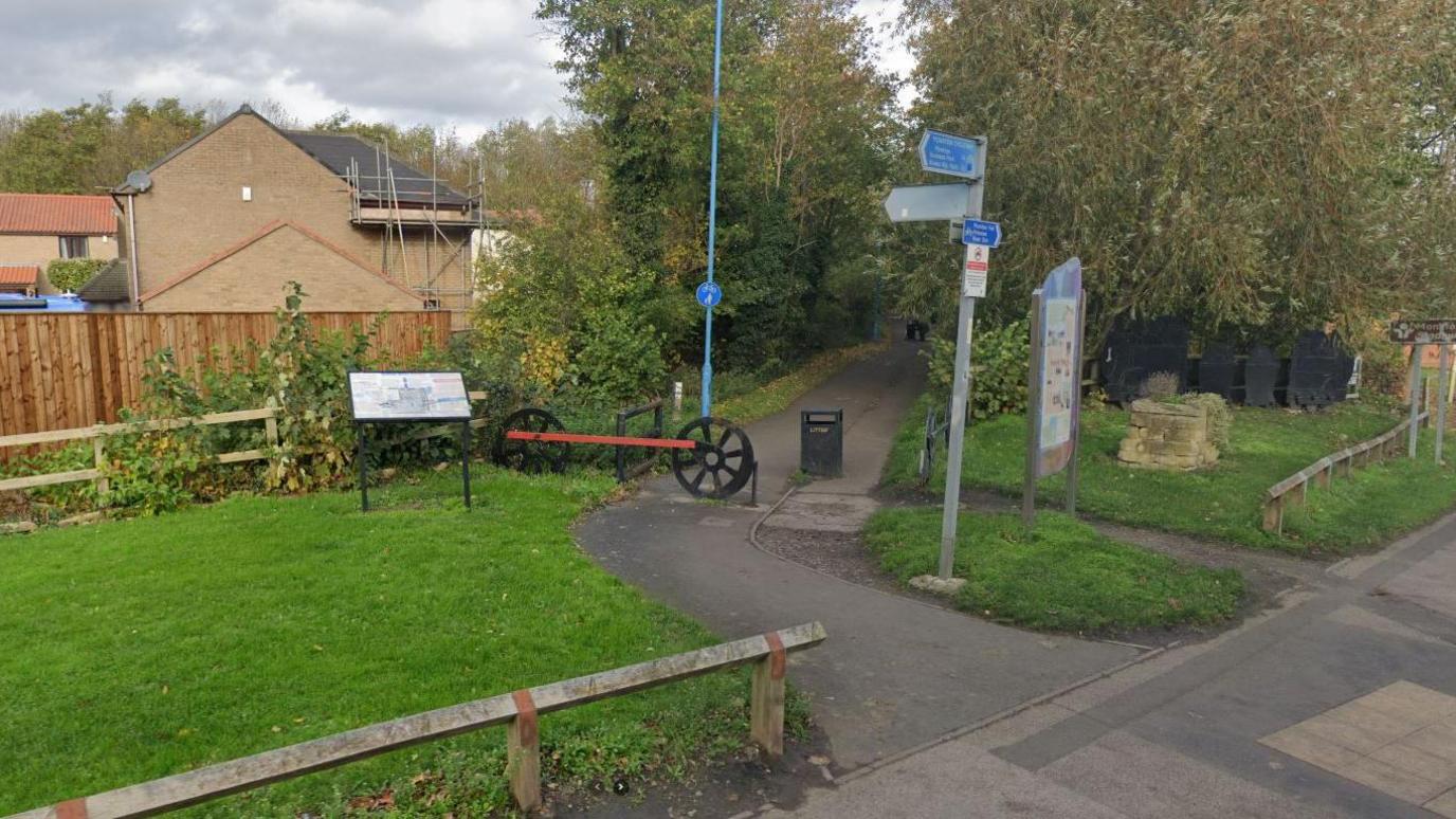 The entrance to Campbell Park in Jarrow. A signpost gives directions to local locations. A sign explains the history of the area. A taller one to the right shows a map of the park. A fence in the shape of train wheels crosses parto f the path leading to the park.