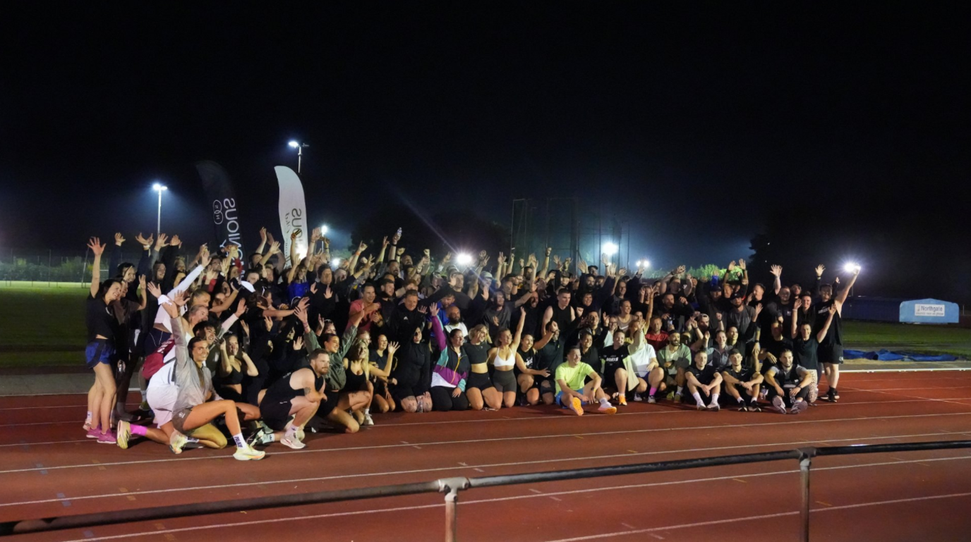 The Harmonious Wellness Club pose together on their track night event at an athletics circuit. There are around a couple of hundred people huddled in a group in their running gear cheering, with their arms in the air. Many of the attendees look to be in their 20s and 30s. The track is lit by floodlights.