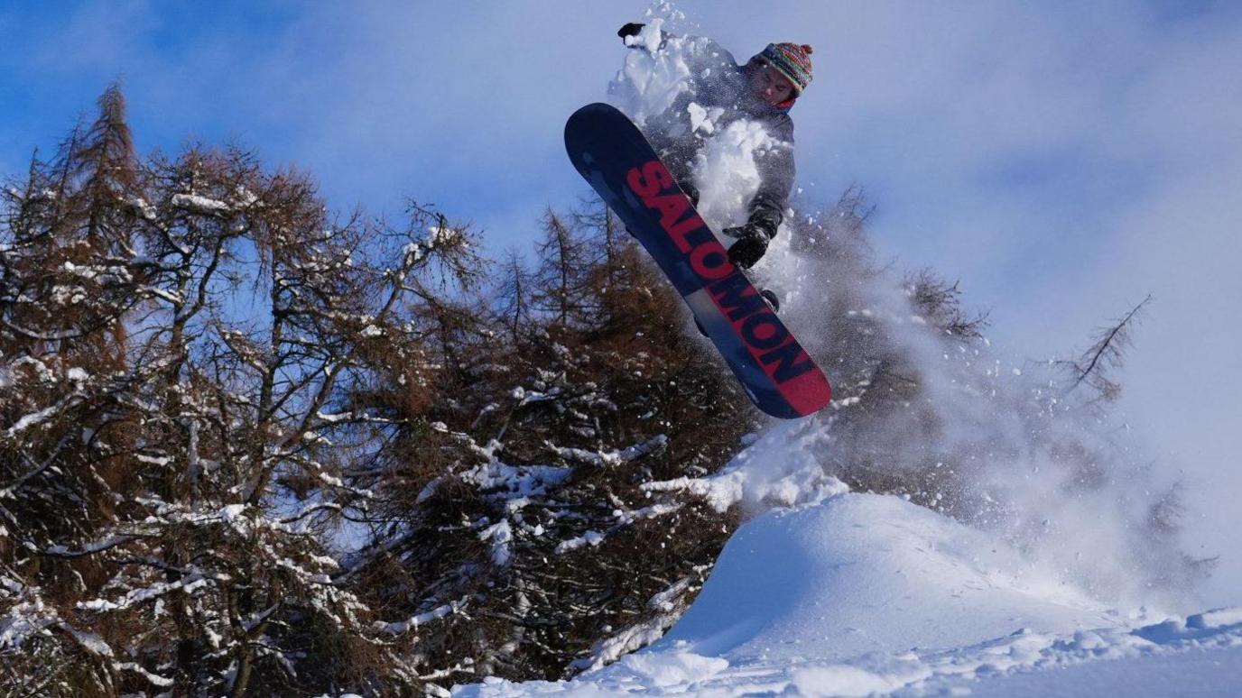 Below view of a snowboarder in the air, jumping over a mound of snow, with a trail of snow behind him. Snow laden trees can be seen to the left, under a clear sky.