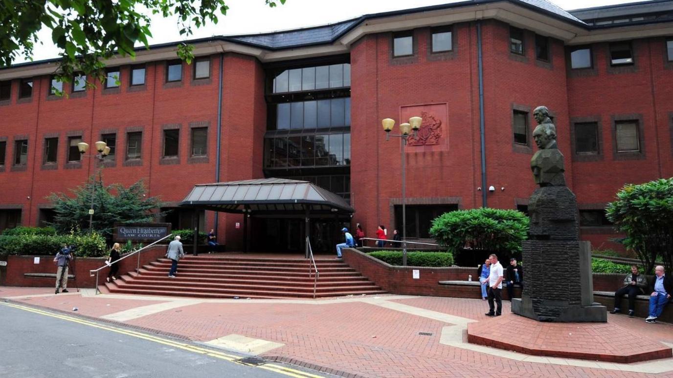 A large, three story-ed red brick building with a canape over the glass front entrance. People are milling about and there is a statyure with four heads on it to the right hand side.