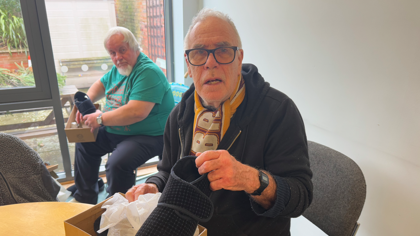 An elderly man wearing glasses and a black hoodie holding a pair of slippers while looking into the camera. 