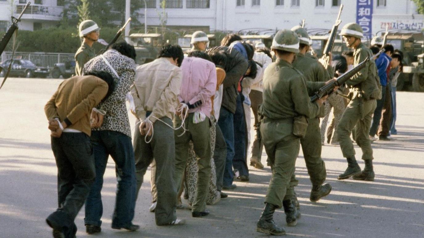 Armed South Korean troops wearing helmets and with large guns round up protestors - mainly men, with their hands tied behind their backs, all in civilian clothes - following a raid in the city of Gwangju after an uprising against the Chun Doo-hwan dictatorship, South Korea, 27 May 1980