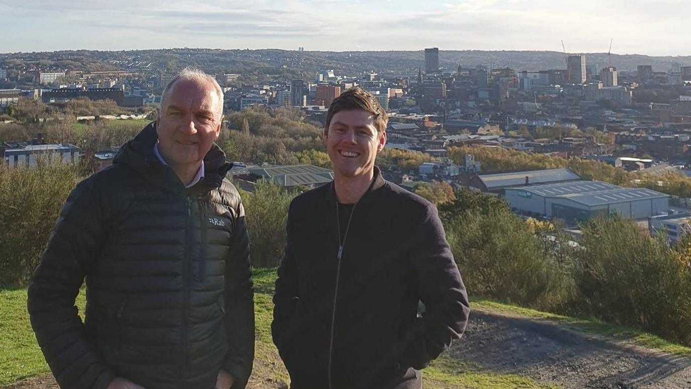 Two men in dark coats stand smiling at the top of a grass bank with a city stretching out below them