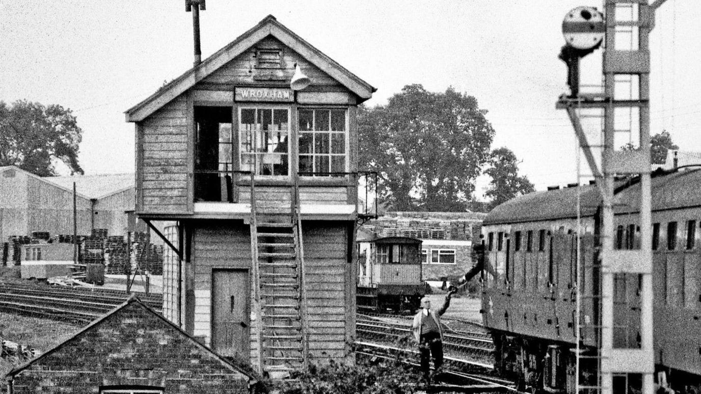 A black and white image of a railway yard with various different train lines and a signal box with steps leading up into it. A railway worker stands next to the railway line passing something to a train driver who leans out of a carriage.