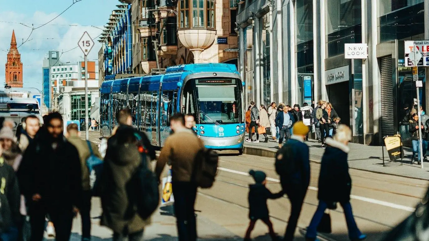A blue tram with people walking in front and behind it in Birmingham city centre.