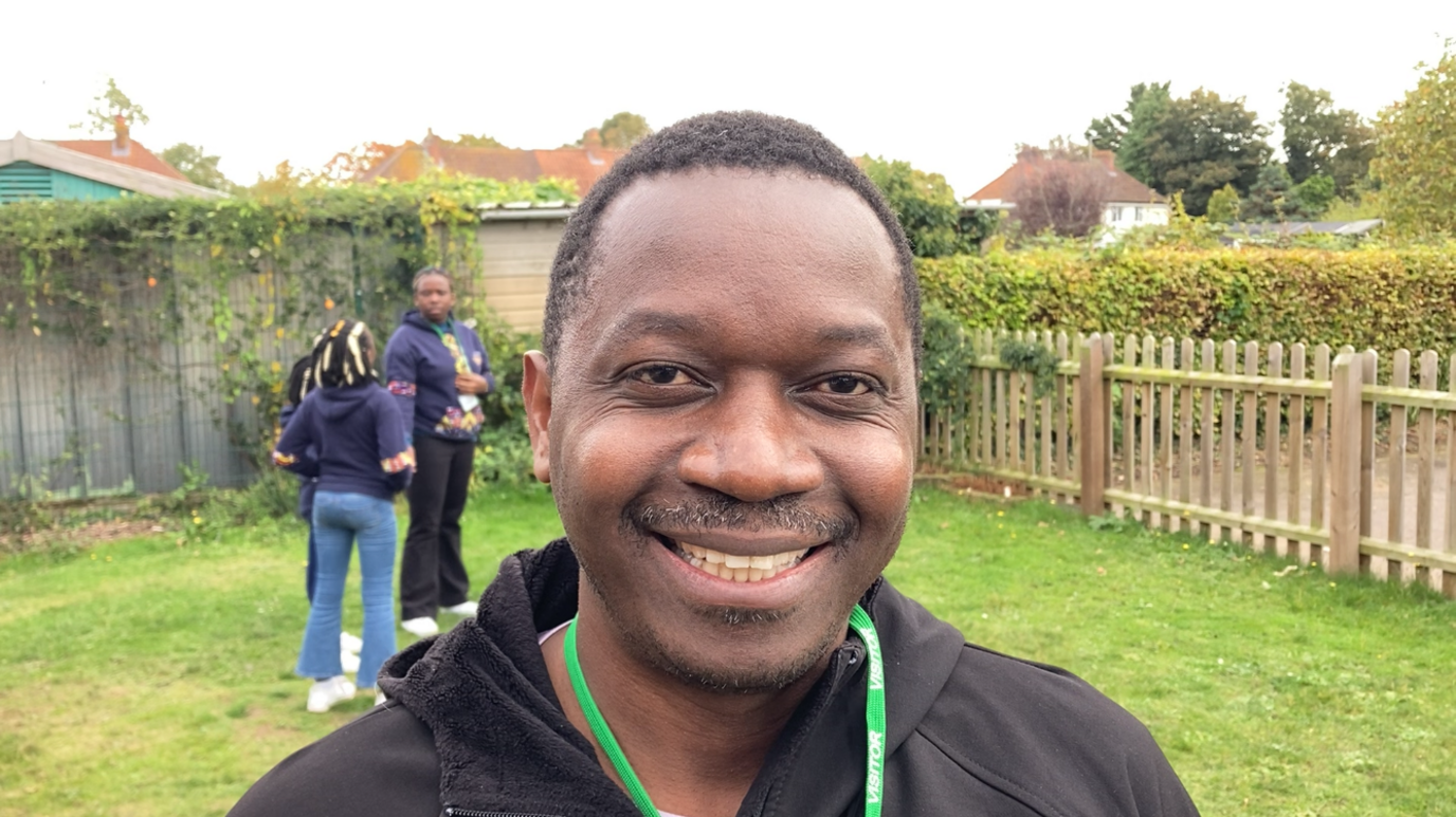 Herbert Mashami is standing outside on some grass and is smiling into the camera. He has short black hair and is wearing a green lanyard. He is wearing a black jacket and behind him are a group of children singing while standing in a circle.