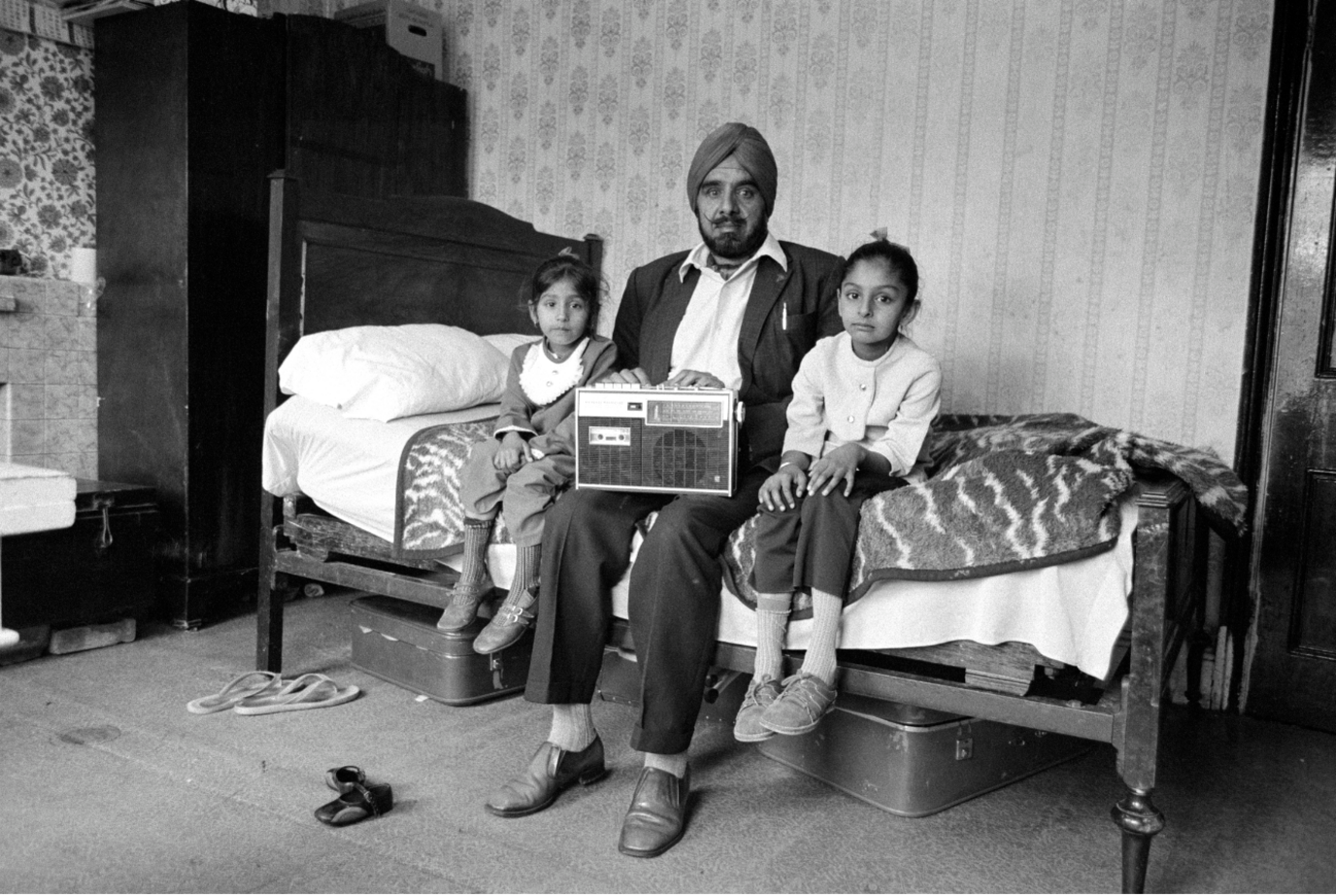 Black and white photograph of a man sitting on a bed with his two small daughters sat on either side of him. A radio set is perched on the man’s lap