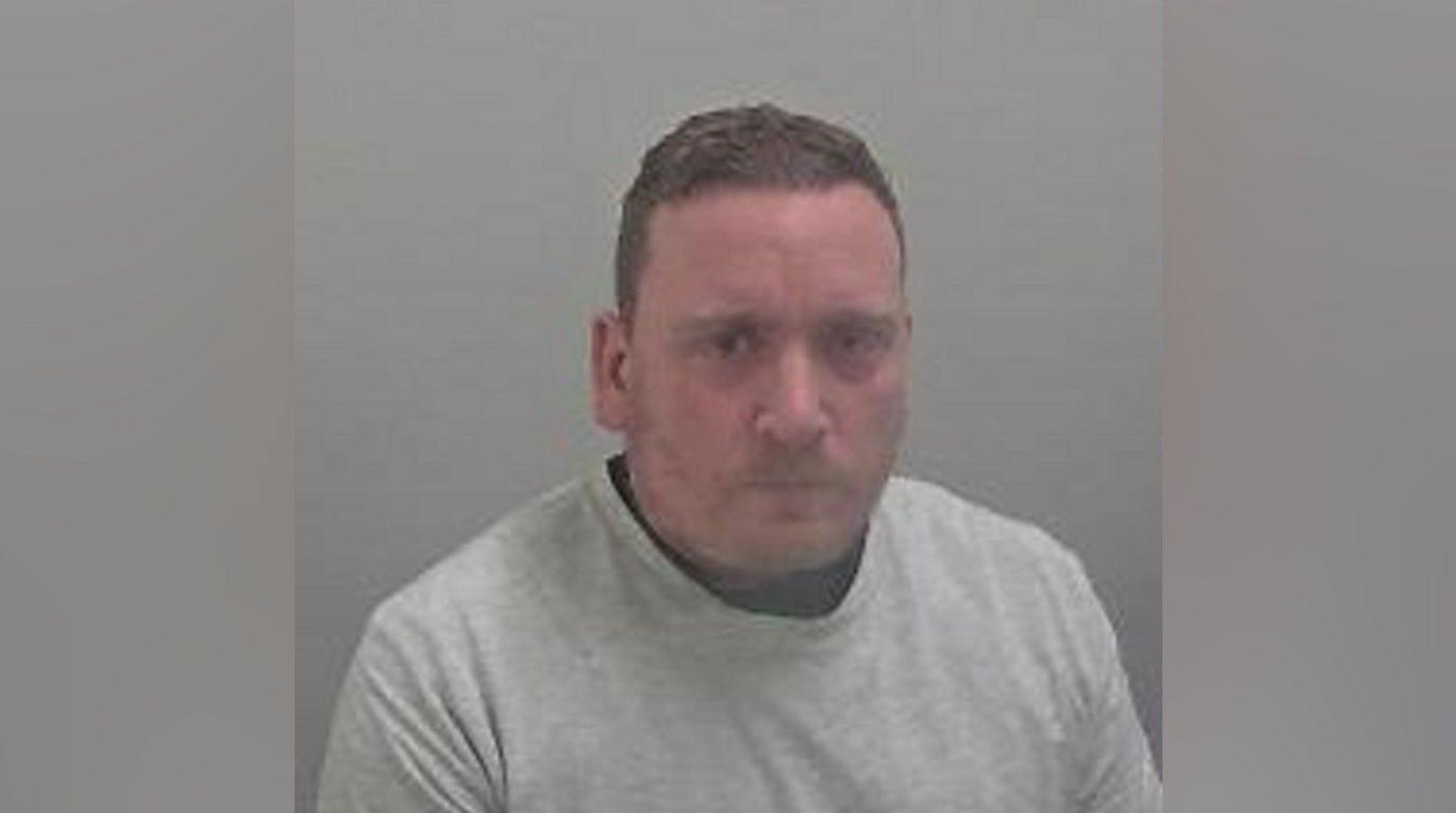 A police custody photo of a man with short brown hair, wearing a grey sweater, looks into the camera.