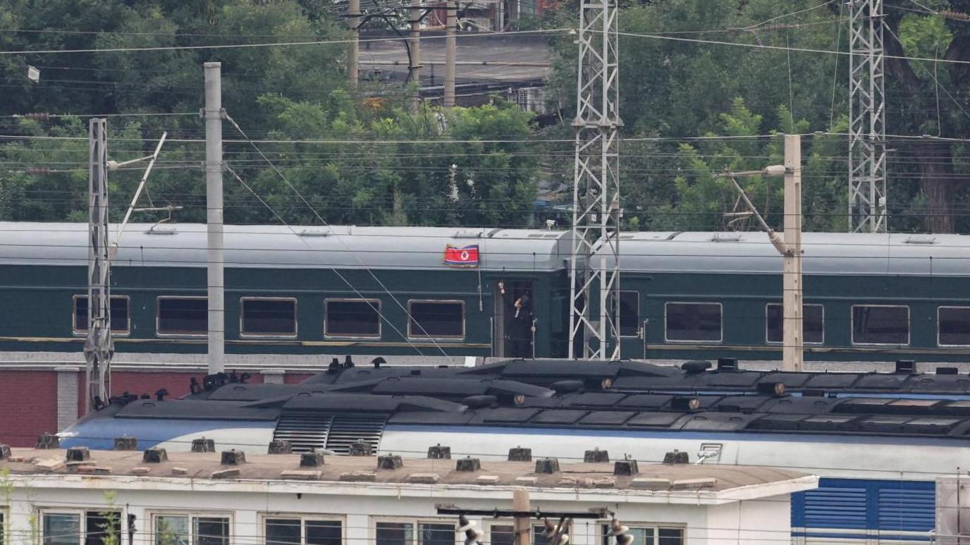 Kim Jong Un's green train is seen going down a railway track with a visible North Korean flag pinned by one of its entrances, while a person in a dark suit hangs out of a doorway and looks up at the flag. In the foreground and background are residential buildings and other trains