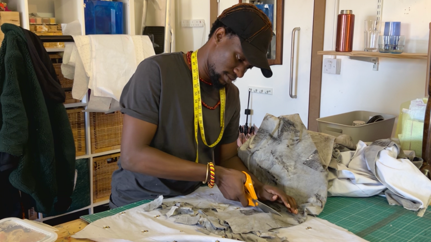 A young, black man, wearing a black cap and a grey T shirt with a red beaded necklace on and a yellow and red striped bracelet. He has a yellow measuring tape wrapped around his neck, and is cutting a piece of grey, tie-dyed fabric with a pair of orange scissors. He is standing at a table with other bits of fabric placed across it.