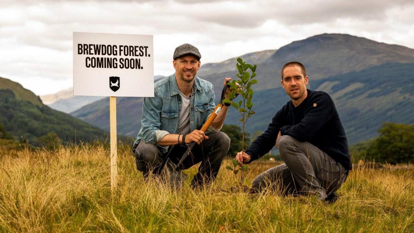 james watt in a cap and blue shirt holding a spade and martin in a long sleeve navy top crouching down in a rural landscape with a sign beside them saying 'brewdog forst coming soon'