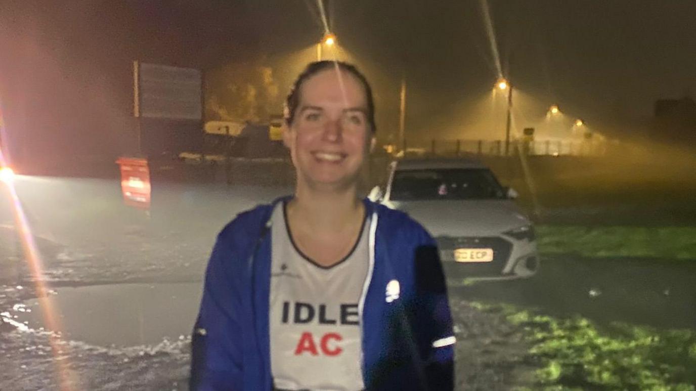 Ellie stands outdoors at night on a wet, uneven surface with patches of grass nearby. She is wearing a blue jacket over a white shirt with the text “IDLE AC” printed on it. Behind her, there is a parked car and several streetlights casting a yellow glow through mist or fog, creating a hazy atmosphere.