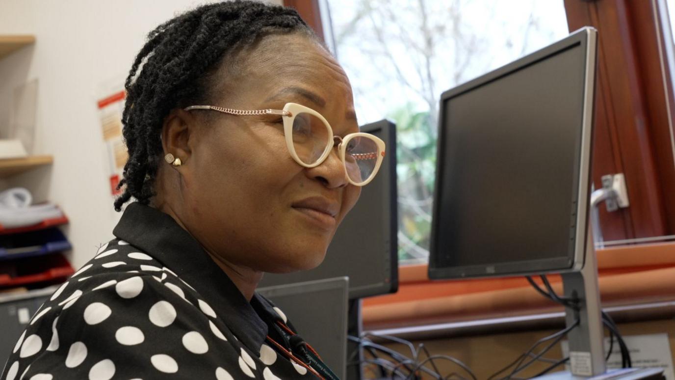 Mosun is seated at a desk in an office setting. She is wearing round glasses and a black and white polka dot top. In the background, computer monitors and a window with a view of greenery.