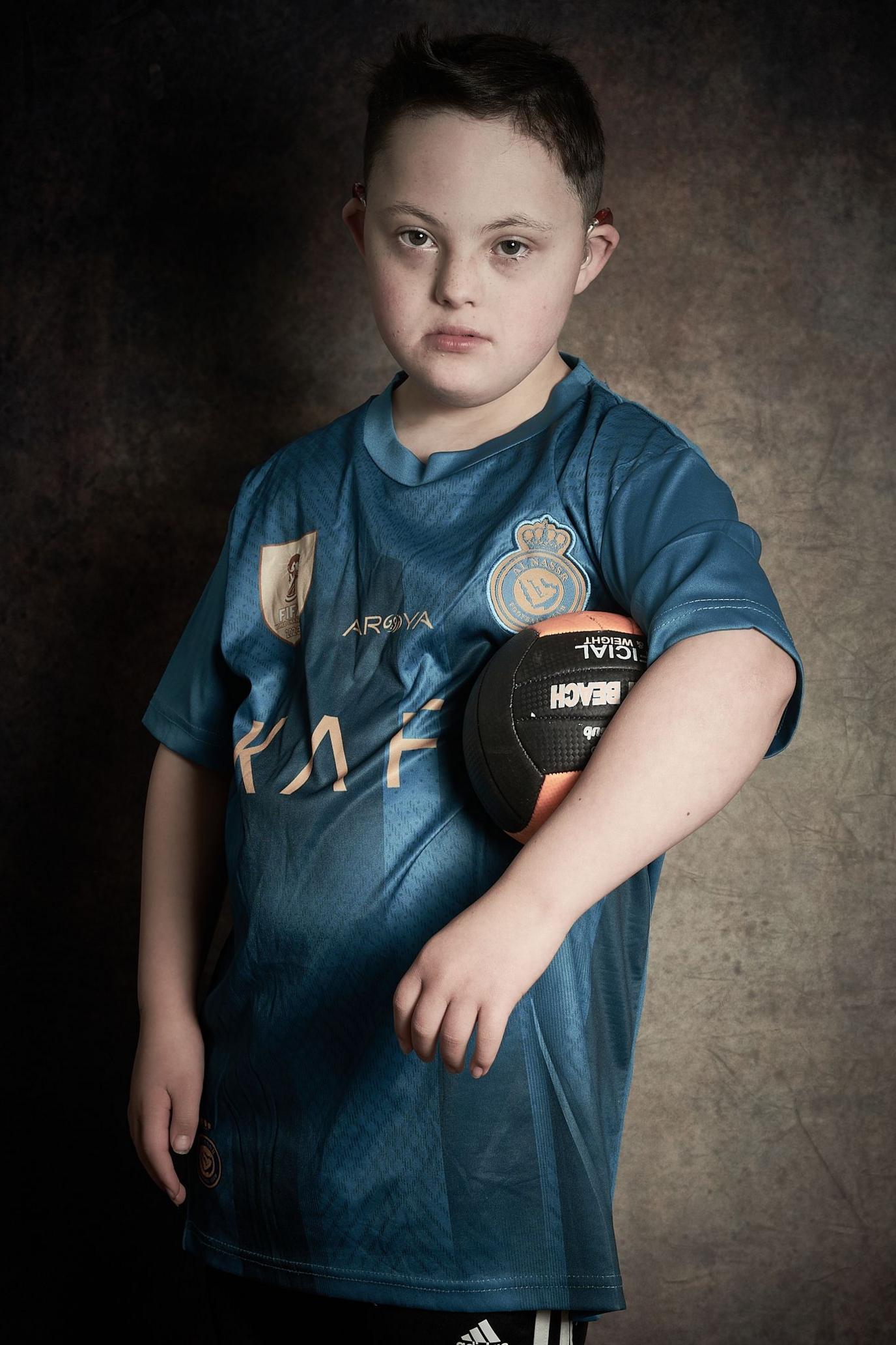 Zack, a boy with short dark hair, looks at the camera. He is wearing a blue football shirt and has a black and orange football tucked under his left arm.