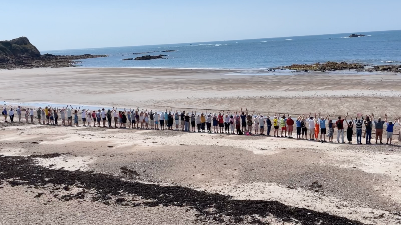 A row of people are seen standing on a beach, their arms held aloft, the sea stretching before them 