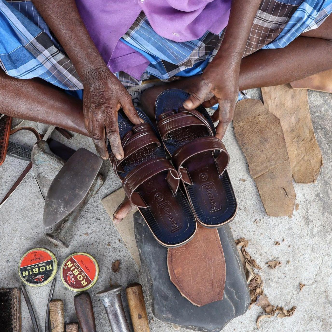 A close-up image of a cobbler's hands holding a pair of maroon leather sandals, surrounded by the tools of his trade