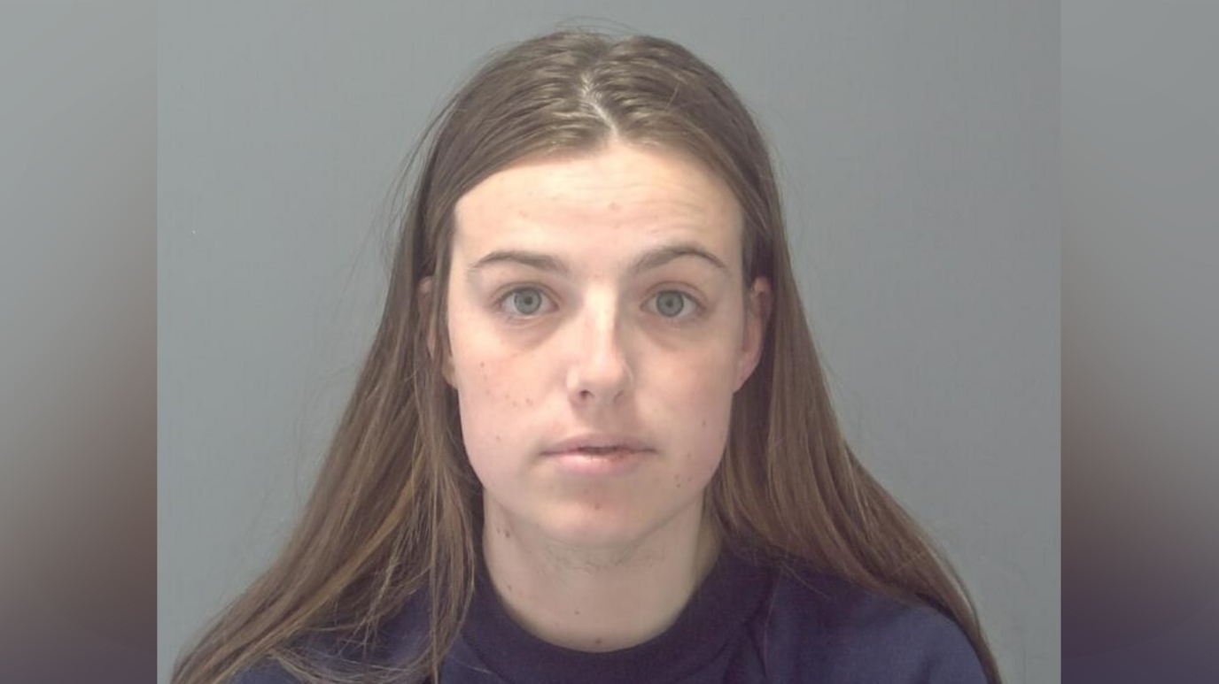 A police custody picture of a woman with long brown hair. She has no expression on her face and is staring at the camera.