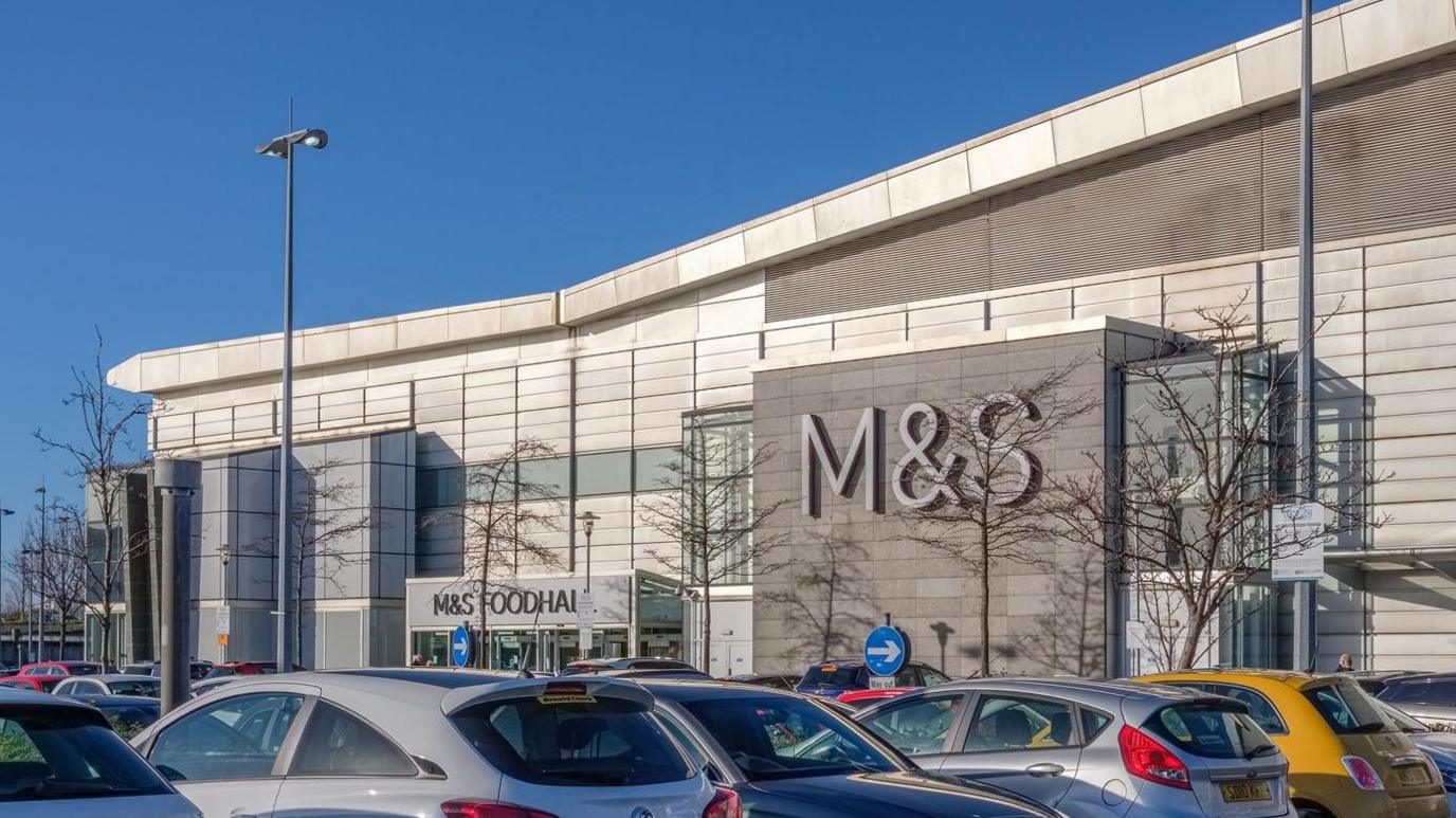 A shopping centre with a sign for M&S with a number of parked cars in the foreground