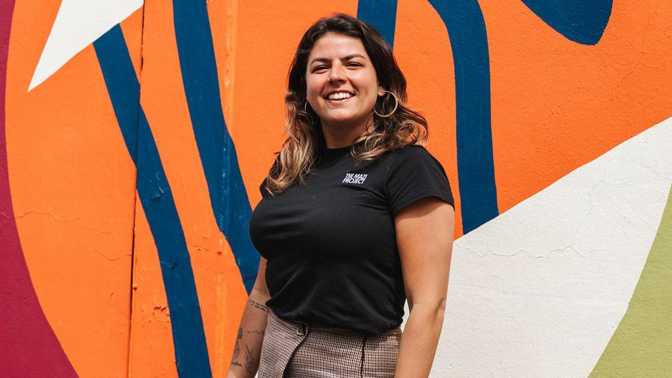 Melanie Vaxevanakis stood in front of a orange and blue mural. She has brown hair and is wearing a black top.