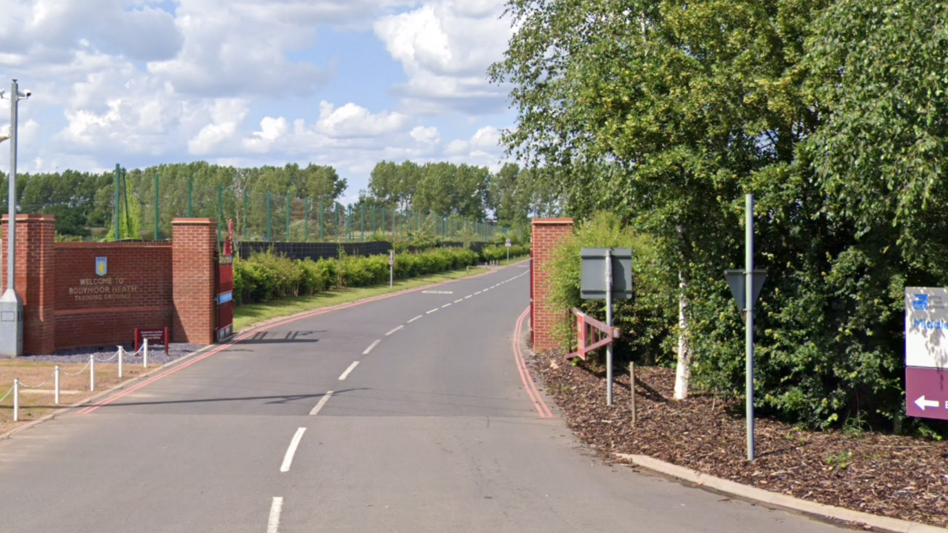 The entrance to the Villa training ground, with a road surrounded by lots of green trees and bushes. A red brick wall is on each side of the road with a Villa logo on the wall