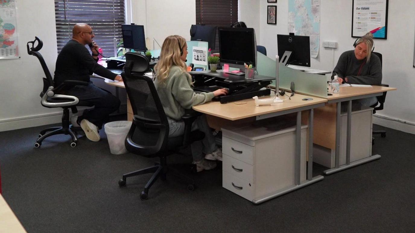 Four employees, looking at screens, sit in an office, two on one side and two behind.