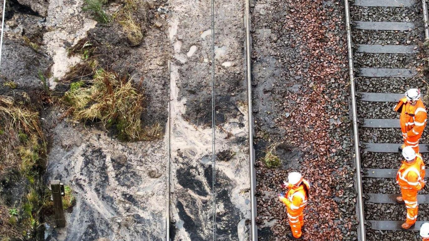 Four workmen in orange hi-vis stand on a railway track as they look at the side of it which appears to show a muddy ditch and debris across one of the tracks.