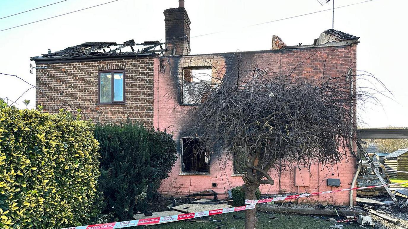 Two semi-detached brick houses badly affected by a fire. The one on the right has pink bricks with the windows burnt out and the roof is missing. There is a burnt tree in front with a red and white cordon tape wrapped around it, stretching across the front lawn. The neighbouring house on the left has brown bricks and its roof is partially missing. The house is covered by two bushes - one has lemon and lime leaves, the other has dark green shrubs