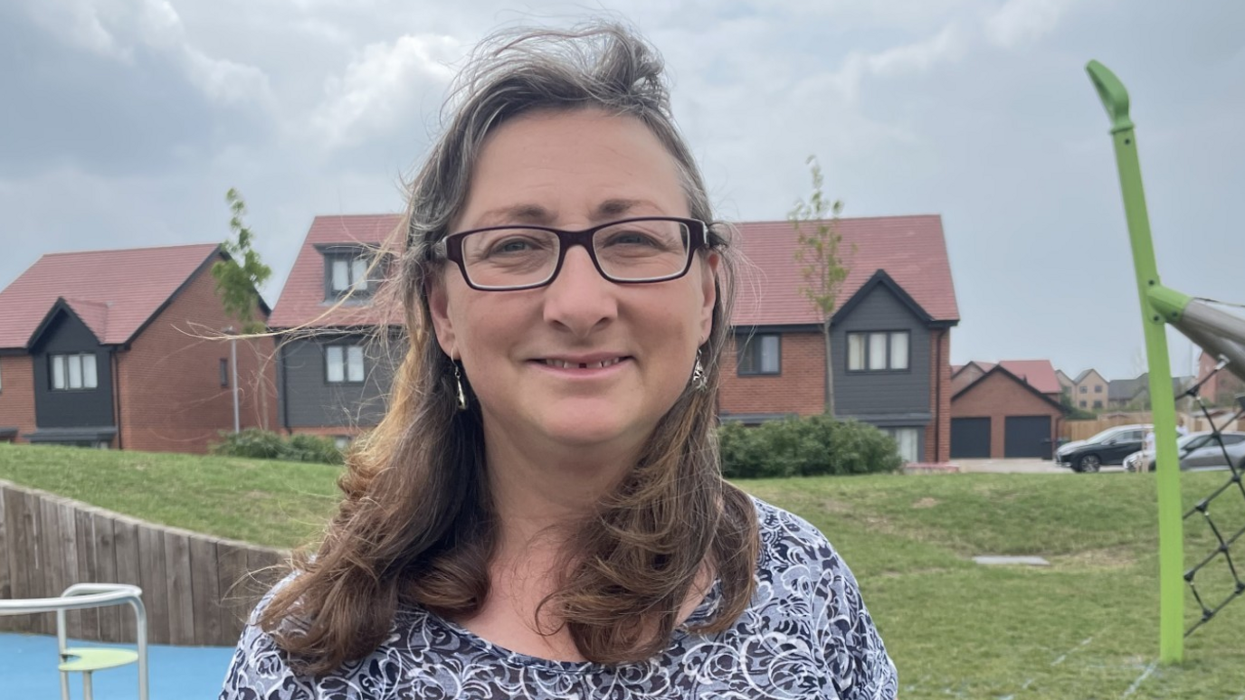 Lindsey is wearing rectangular glasses and has shoulder length hair. She is standing in a playground in front of some new houses, with a roundabout and a goal net either side of her in the background