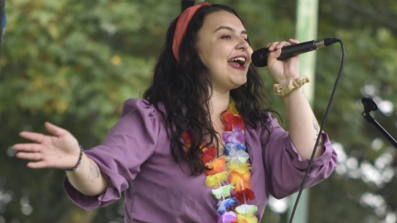 A woman with long black hair and wearing a pink head band, a purple shirt and flowers around her neck, sings into a microphone on stage.