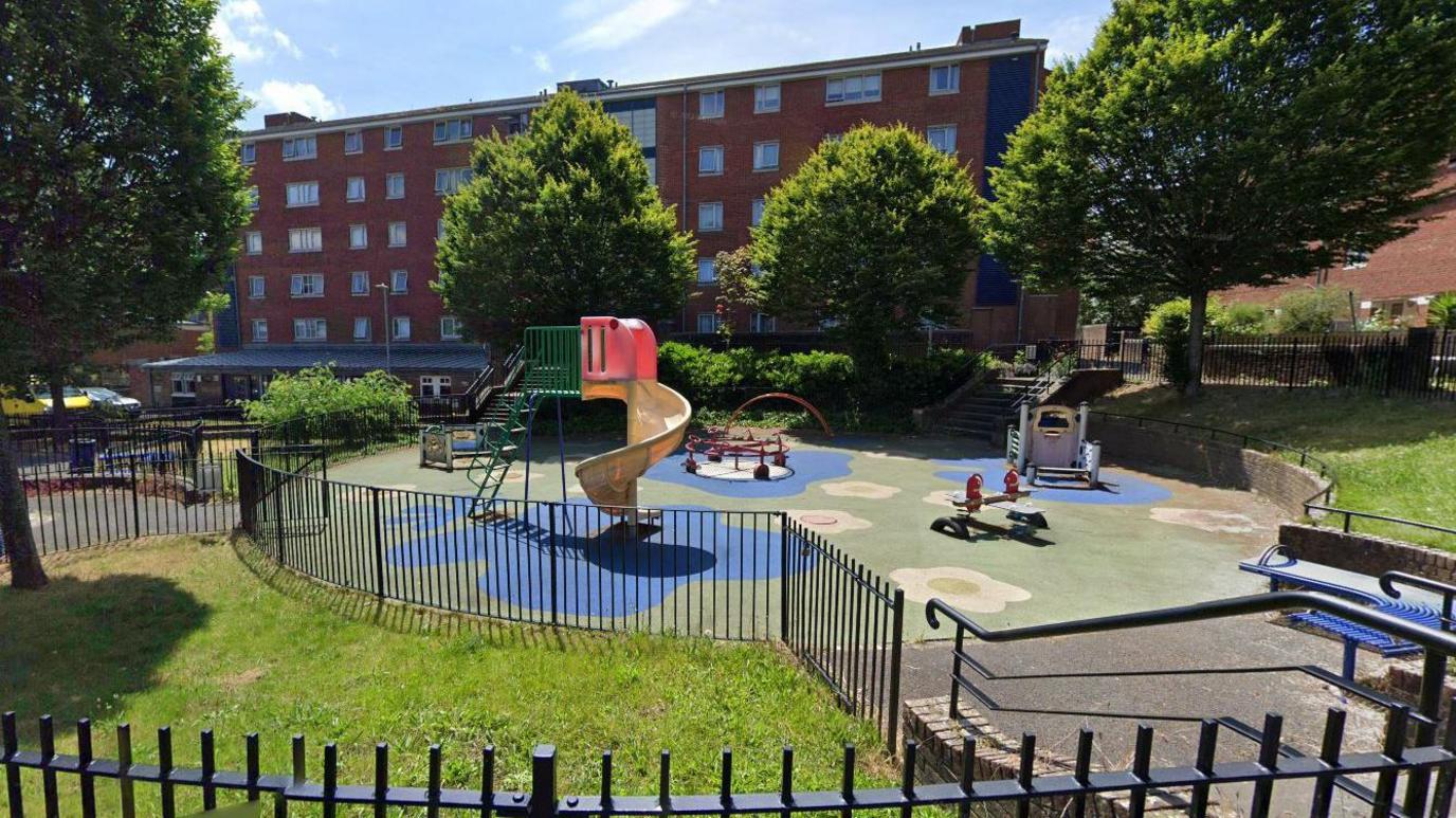 A screenshot from Google street view showing a small children's playpark. There are colourful shapes resembling flowers on the ground, and black metal railings surrounding the perimeter. Inside the park there is a small see-saw, a yellow spiral slide, blue metal benches and several climbing frames. The park is surrounded by bushy trees and in the background there is a brown brick block of flats.