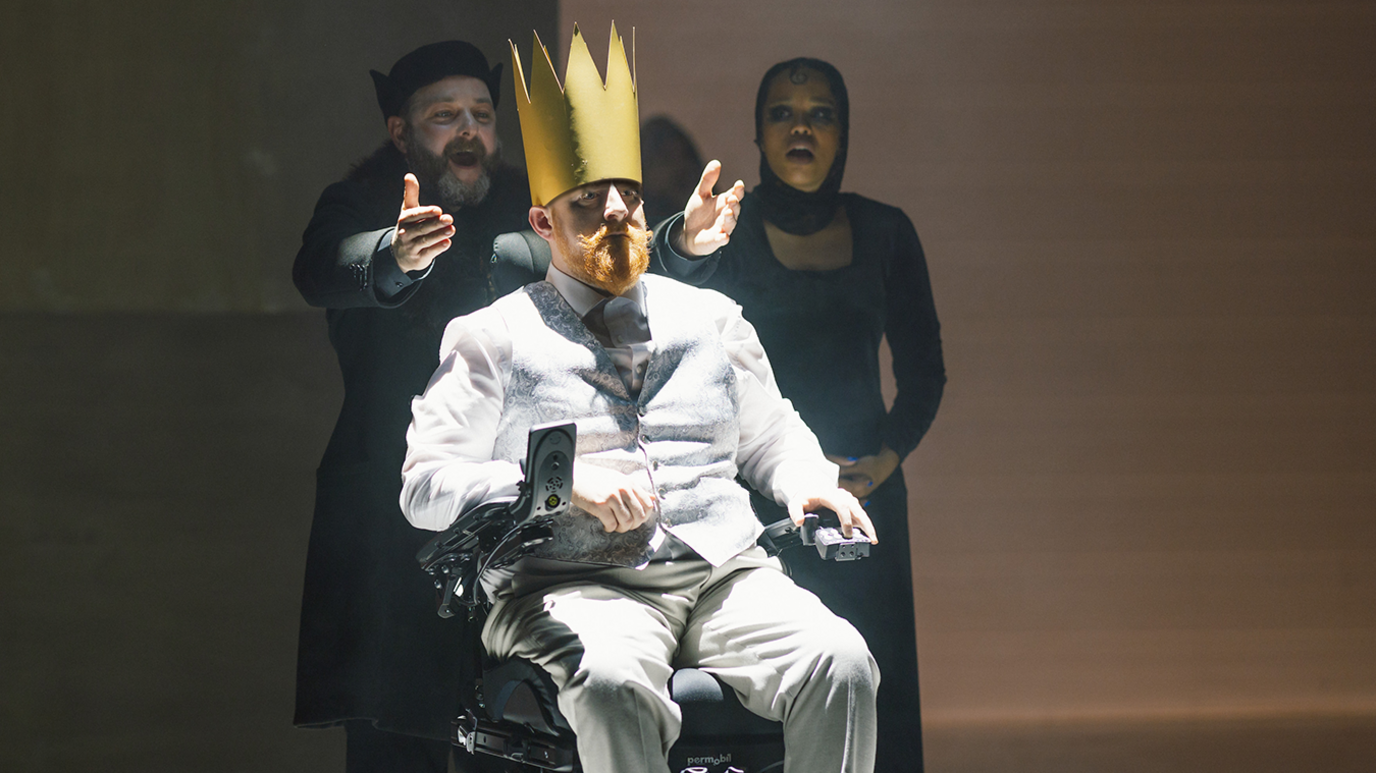 Michael Campbell is pictured in a wheelchair on stage. He is wearing a white suit and has a long golden crown on his head. Two other performers can be seen in the background dressed in all black. 