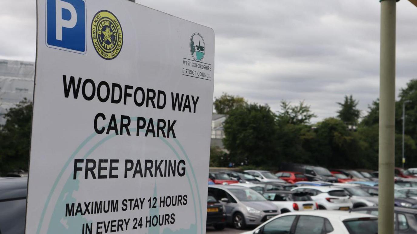 A view of full Woodford Way car park on a cloudy day. A sign for it reads "Free parking. Maximum stay 12 hours in every 24 hours."