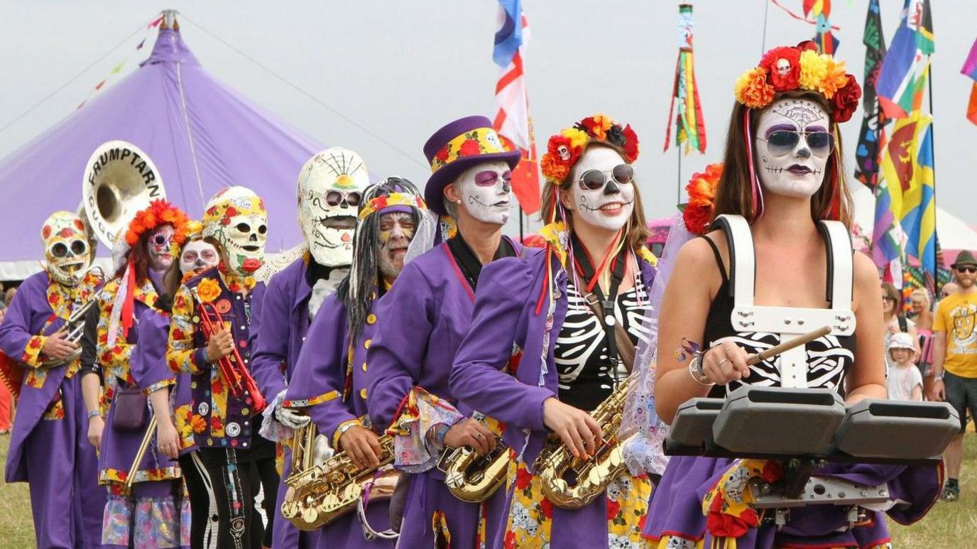 Nine people dressed in purple, stood in a line and holding brass instruments. On their faces are either masks or Day-of-the-Dead style white skull make-up. Some of them have floral headbands on their heads. In the background is a large purple festival tent.