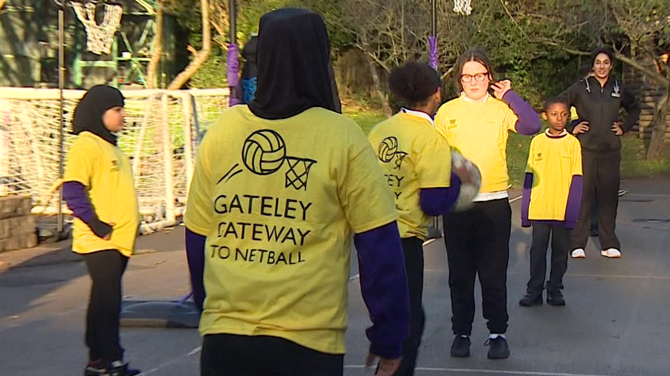 Half a dozen pupils stand on a netball court playing the sport, passing a ball between them. They have yellow T'shirts on that say Gateley Gateway to Netball on the back. An older woman stand behind them watching them.