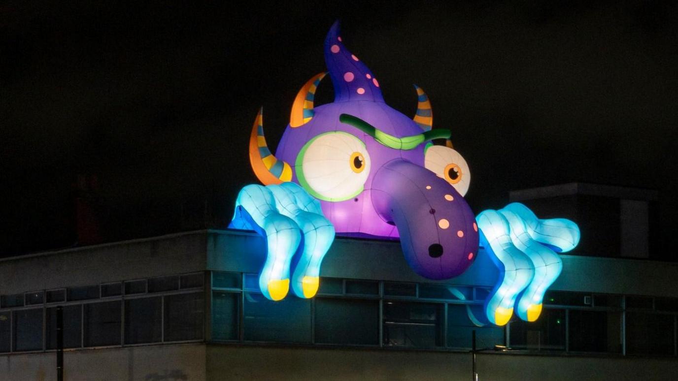 An inflatable lit-up monster with a purple head, long nose and blue hands with yellow fingernails. It has big white eyes and orange and blue spikes coming out its head. It is peeking over the top of a building.