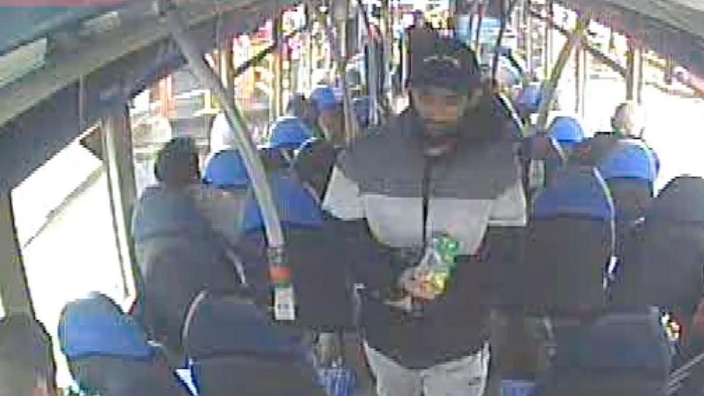 Man wearing a striped puffa-style jacket and a baseball cap walks down the aisle of a bus