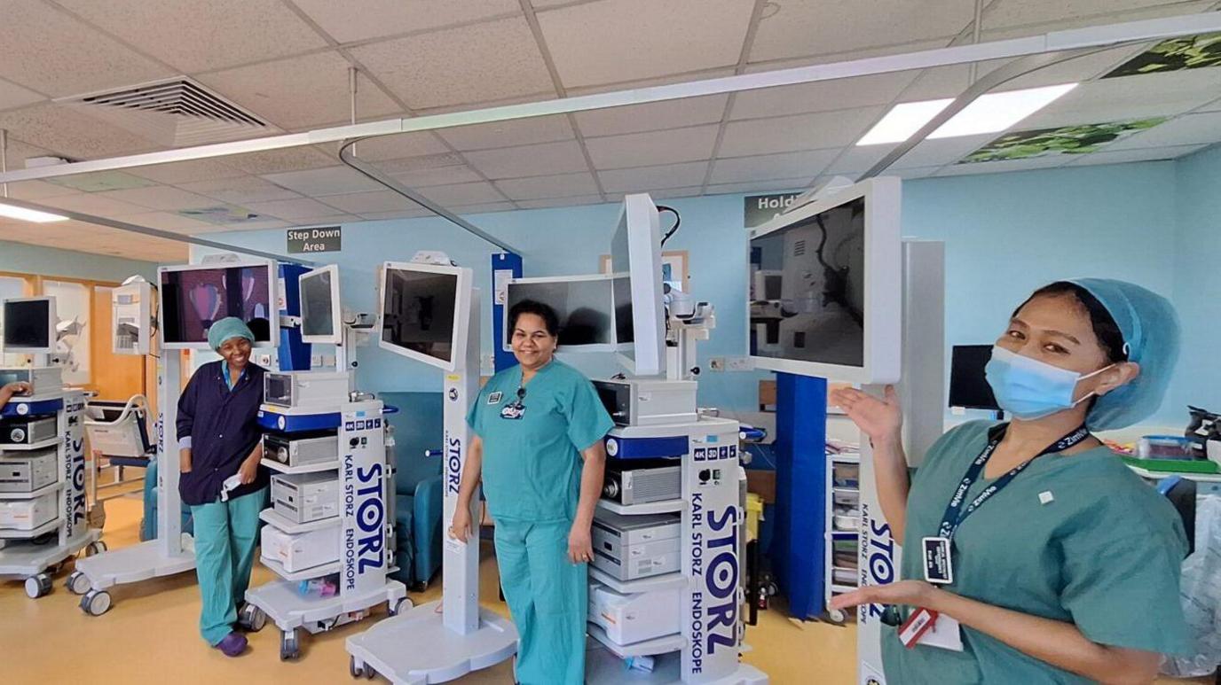 Three member of female hospital staff stand amongst medical equipment. They are all wearing hospital scrubs. The staff member on the right is wearing a face mask - and is gesturing towards the new equipment which has been funded by the charity. 