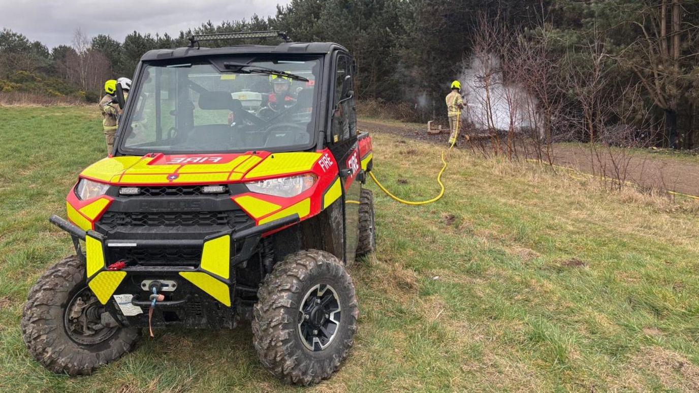 A small fire service vehicle that looks like a mini truck with a pump attached to the back which is being used to put out a fire in the background of the image. A firefighter can be seen holding the yellow tube attached to the pump to mist a pile of discarded batteries near woodland. 