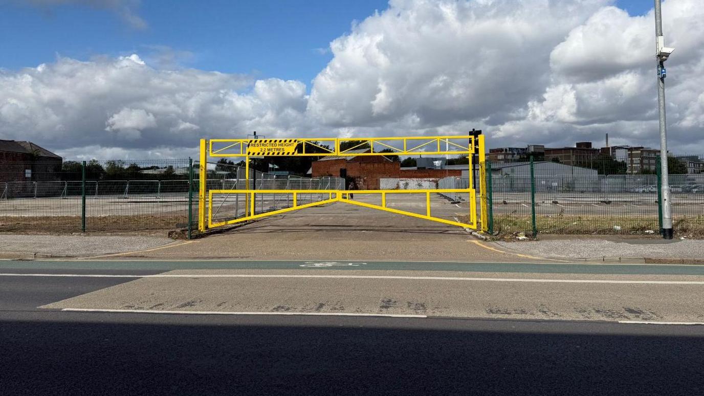 The photo shows a yellow gate which is on the other side of the road. There are green fences either side of it, and an empty car park behind.