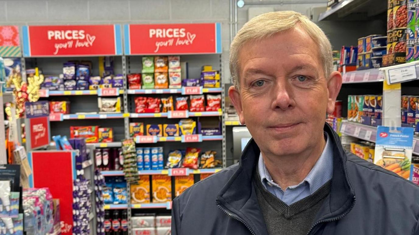 A head and shoulders view of security manager Mark Foulds, who is standing in front of supermarket shelves full of goods and signs reading "prices you'll love". He has short grey hair and wears a light blue shirt, a dark grey pullover and a dark grey jacket.