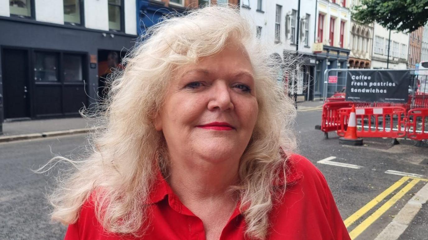 A woman, who has long light blonde hair, wearing a red shirt. She is standing in front of a road in Belfast city centre. 