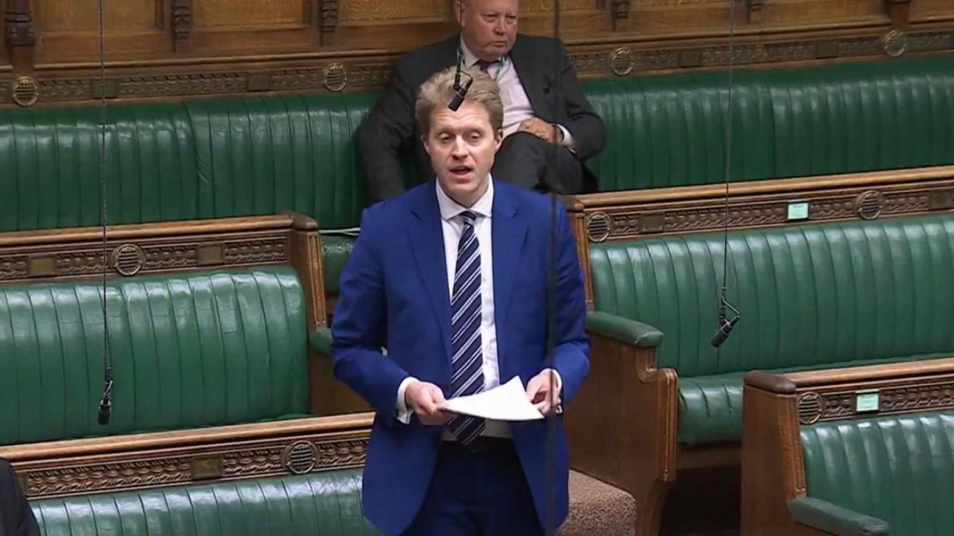 Dr Ben Spencer, the Conservative MP for Runnymede and Weybridge, speaking in the House of Commons. He is wearing a blue suit and a striped tie. The green benches of the Commons and another MP can be seen behind him.