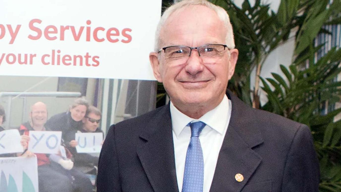 A smiling Tom Flood, wearing a navy blue suit, white shirt and pale blue tie. He has white hair and is wearing glasses.