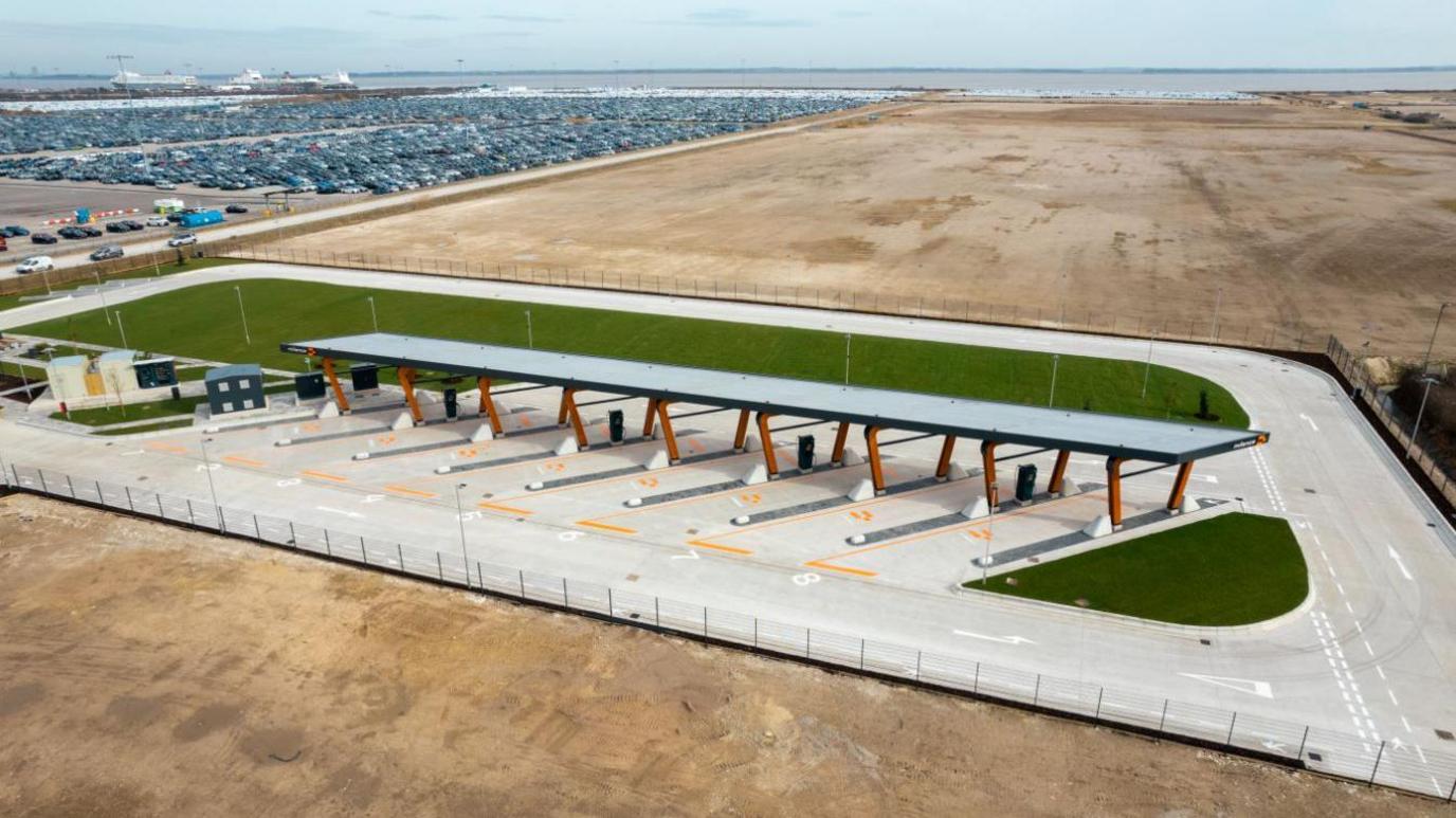 An aerial adaptation of what the design of the HGV electric charging point would look like. A roof can be seen over eight charging ports with grass behind and a large car park next to it.