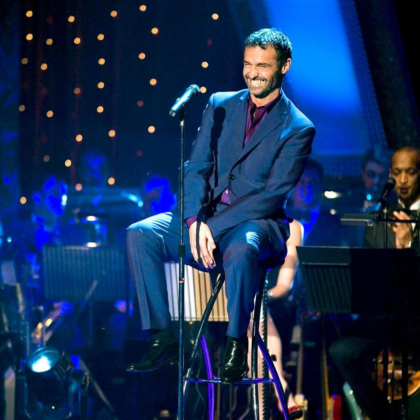 An image of Pellow on the set of Strictly - sitting on a stool in front of the live orchestra. He is in a dark blue suit and leans back, receiving applause and smiling back at the audience.