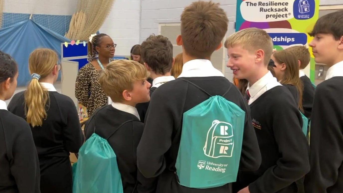 School pupils in black jumpers with white shirts and red ties, they are stood in a school hall talking and some have green backpacks on their backs which have university of reading logos on them and a resilience rucksack symbol.