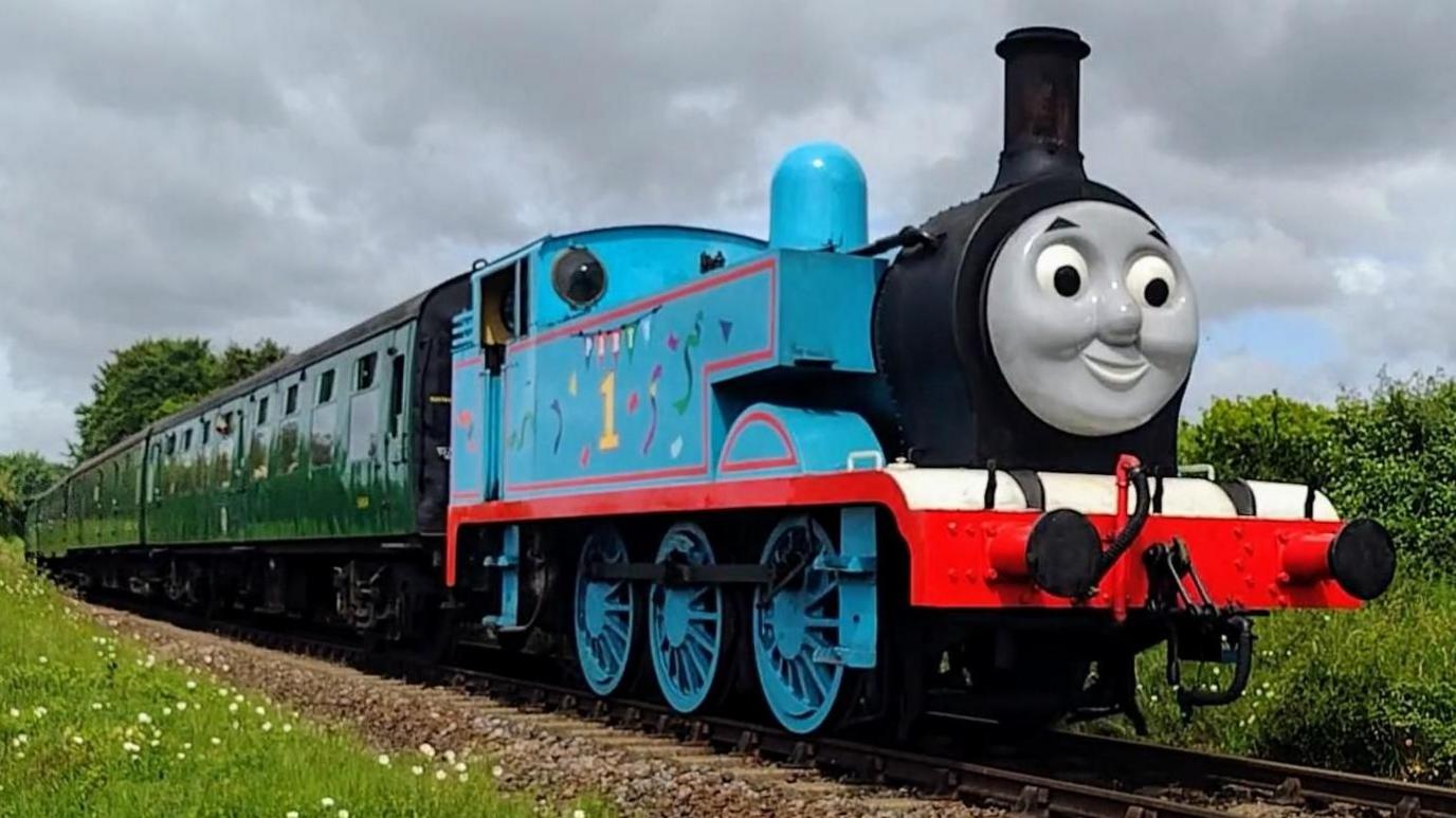A life size Thomas the Tank Engine on a train track. Thomas is a mainly blue steam engine with a happy face, pulling green coaches behind him.