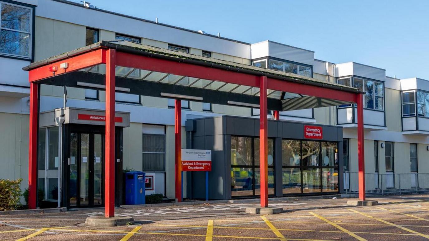 The front of the Queen Elizabeth Hospital in King's Lynn. The foreground shows the yellow hatched area in front of the accident and emergency department. The entrance has a red metal frame and the background shows the aged two storey building, built with crumbling Raac concrete