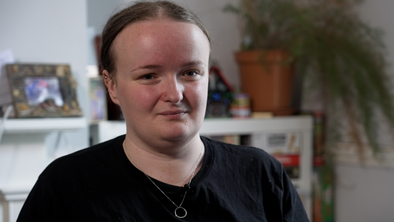 Lily has her brown hair tied up and is looking behind the camera. She's wearing a black T-shirt and a silver necklace. Sat in front of a homely background with a photo frame and plant.