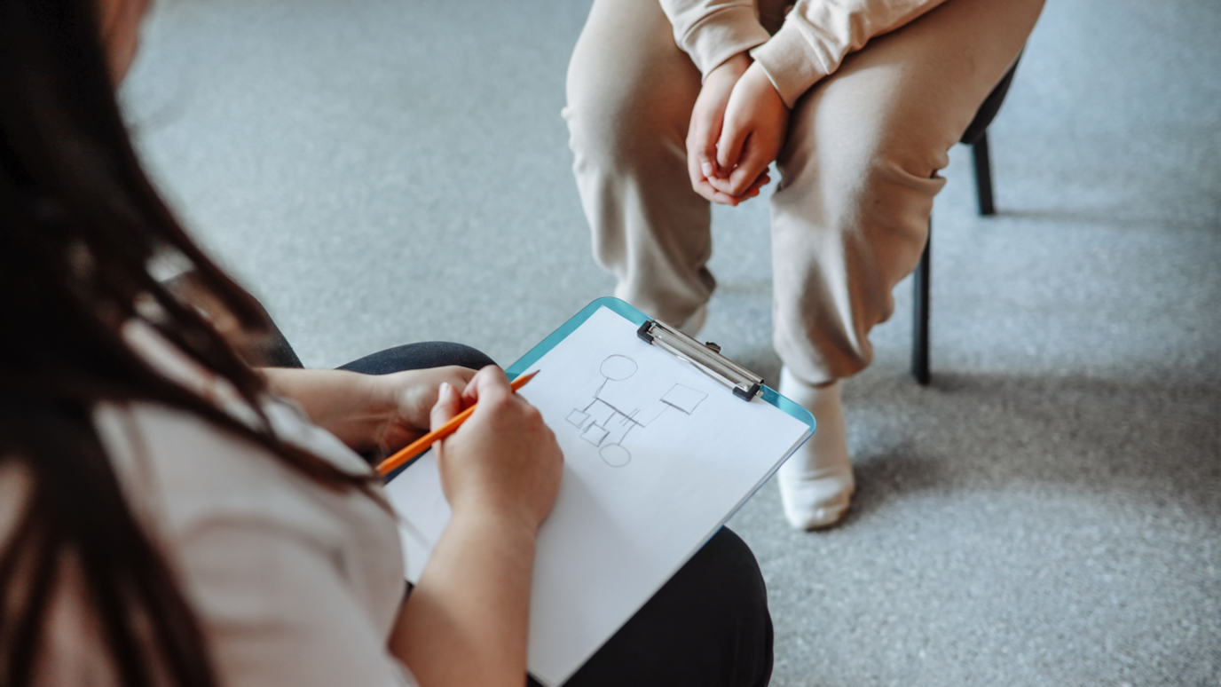 A child sits on a chair with a drawing on a clipboard. A second person's legs are visible. They are sitting on a chair facing the child. 